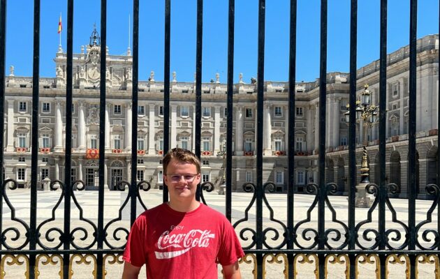 Kaleb leaning on a fence in front of a large building