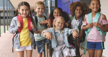 Diverse group of children standing with a peer who is using a wheelchair, smiling and wearing backbacks